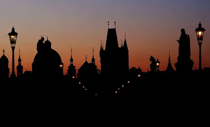 Silhouettes of bridge statues and towers against an orange sky in Prague.